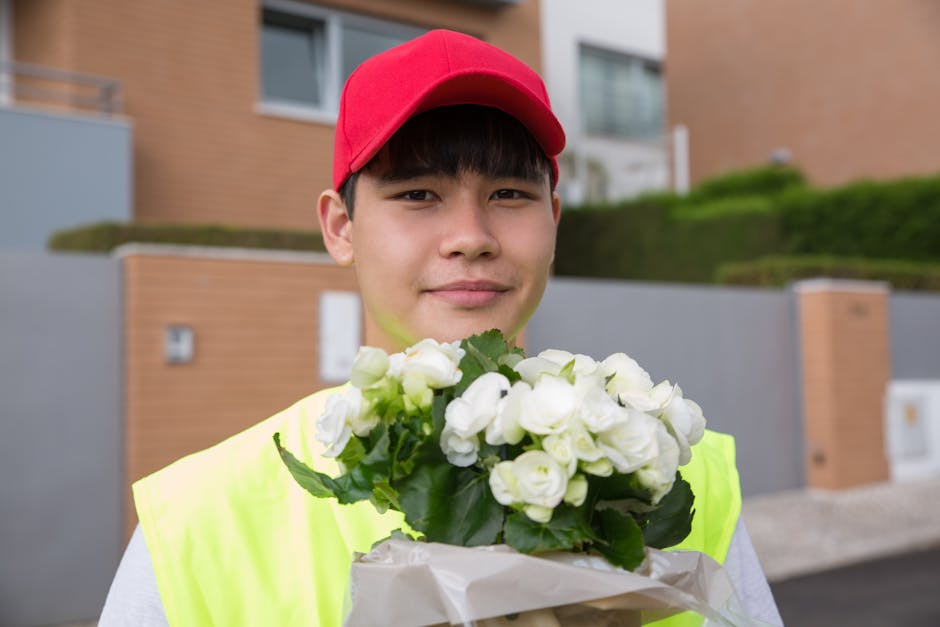 A young delivery person wearing a bright yellow high-visibility vest and a red baseball cap is holding a bouquet of fresh white flowers wrapped in white paper, standing outdoors in front of modern res