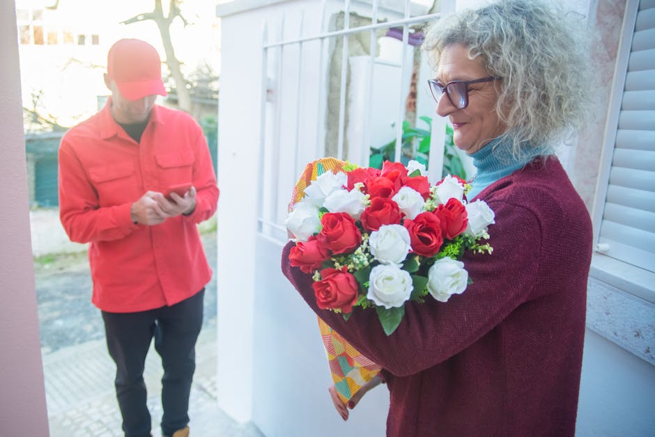 A woman with curly gray hair and glasses, dressed in a maroon sweater and blue turtleneck, is holding a vibrant bouquet of fresh roses in red and white with green foliage, wrapped in patterned paper.