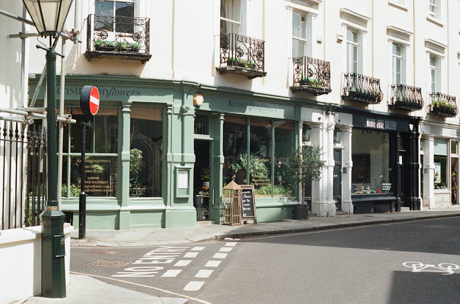A charming flower shop on Kingsland Road Haggerston with a pastel green storefront, large display windows showcasing fresh flowers in various colors, and potted plants placed outside. The shop has dec