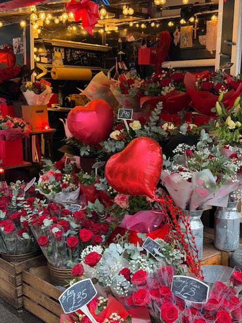 A vibrant display of fresh red roses and white flowers arranged in bouquets at a flower stall, with two large red heart-shaped balloons among the floral arrangements. The flowers are wrapped in clear 