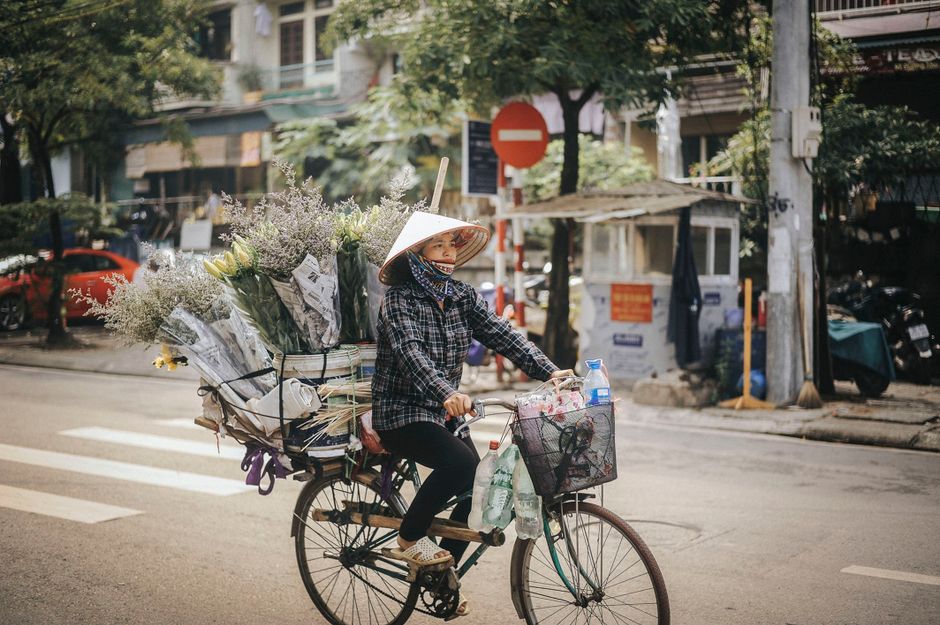 A woman cycling on a city street, carrying a large load of flower bouquets wrapped in newspaper and transparent plastic, featuring various blooms like yellow roses, white baby's breath, and purple-tin