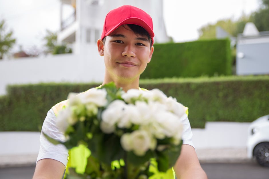 A delivery person wearing a bright yellow vest and a red cap holding a bouquet of white flowers with green leaves, presenting it towards the camera outdoors on a residential street with a white buildi